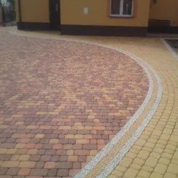 Zakrzywiony podjazd wyłożony kostką brukową w odcieniach brązu i żółci, z jasnoszarą obwódką, prowadzący do budynku z żółtą elewacją.