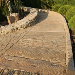 Zakrzywiona ścieżka w ogrodzie, wykonana z desek imitujących drewno, obramowana kostką i kamiennym murkiem, otoczona zielenią.