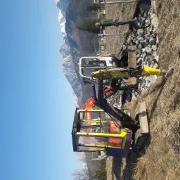 Dwa minikoparki na tle górskim i błękitnego nieba, jedna z żółtym młotem hydraulicznym, obok leży rura, a w tle pracownik zabezpiecza teren.