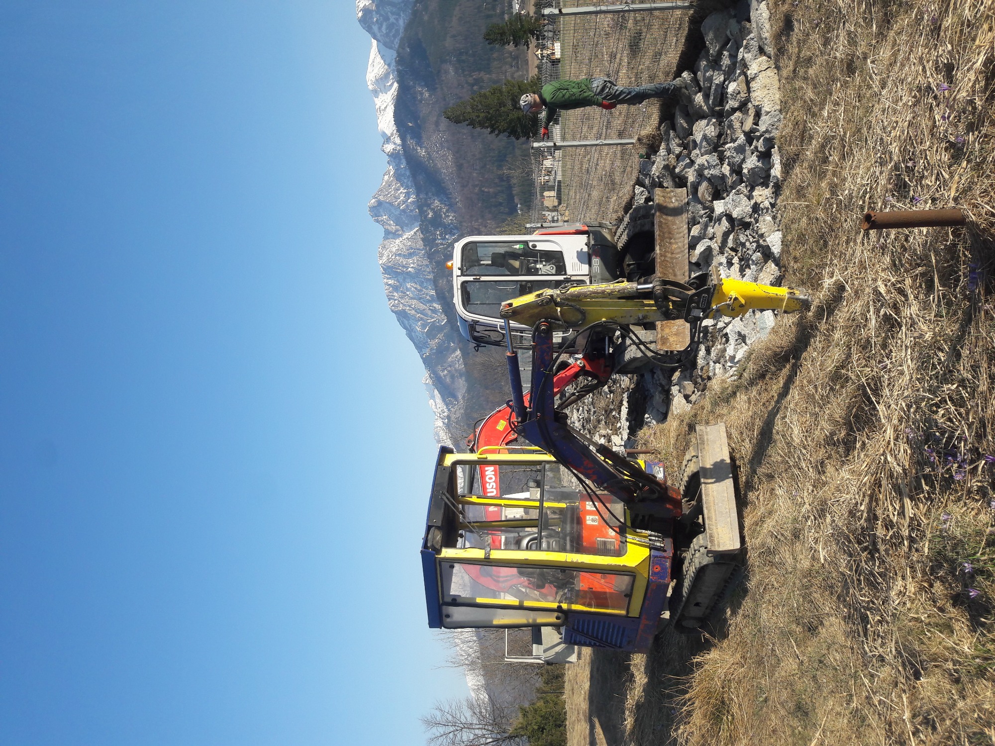 Dwa minikoparki na tle górskim i błękitnego nieba, jedna z żółtym młotem hydraulicznym, obok leży rura, a w tle pracownik zabezpiecza teren.
