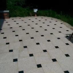 Wyłożony jasnoszarą kostką taras z regularnie rozmieszczonymi czarnymi wstawkami, z drewnianymi krzesłami ogrodowymi po prawej stronie i zielenią w tle.