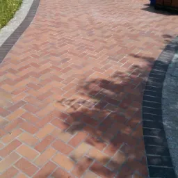 Ułożona z ceglastych kostek ścieżka w jodełkę, obramowana ciemniejszymi kostkami i szarym kamieniem, z paletą kostek w tle i cieniem drzewa na powierzchni.