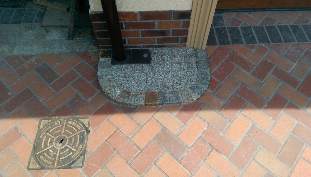 Fragment chodnika z ceglanej kostki ułożonej w jodełkę, z półokrągłym obramowaniem z szarej kostki granitowej przy rurze spustowej, studzienka kanalizacyjna w rogu.