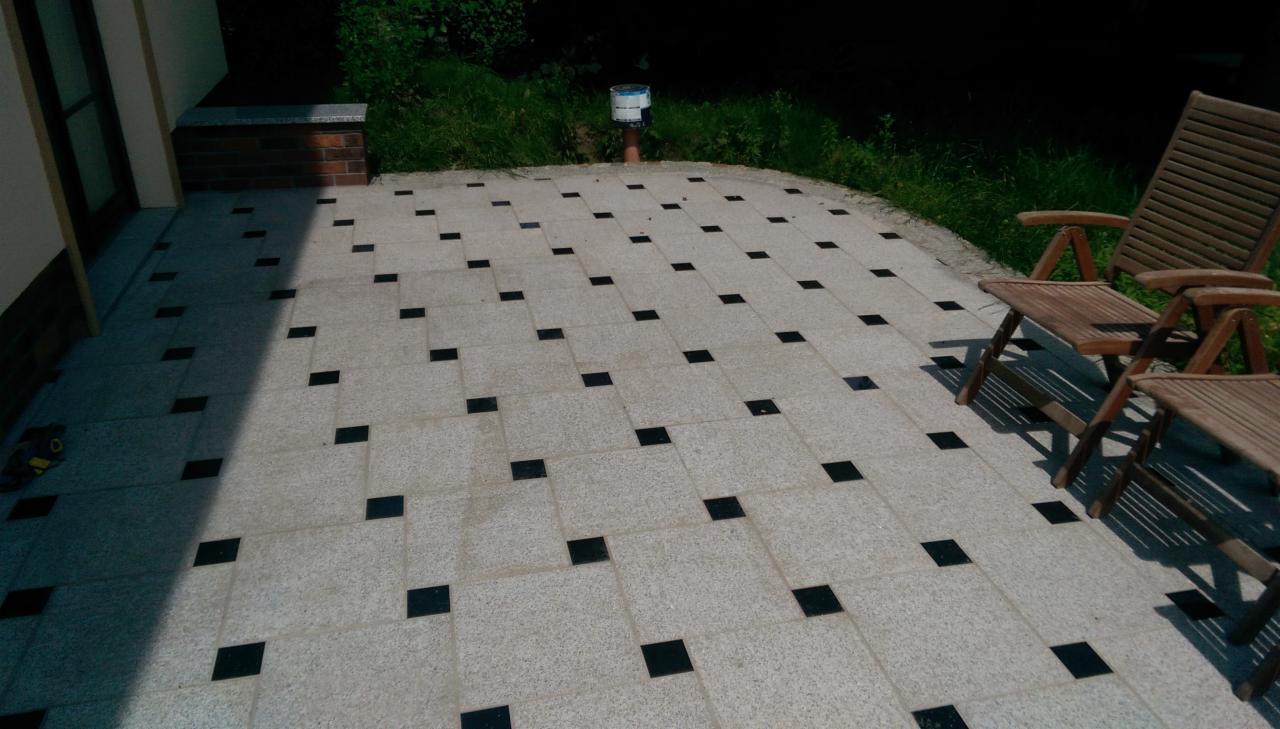 Wyłożony jasnoszarą kostką taras z regularnie rozmieszczonymi czarnymi wstawkami, z drewnianymi krzesłami ogrodowymi po prawej stronie i zielenią w tle.