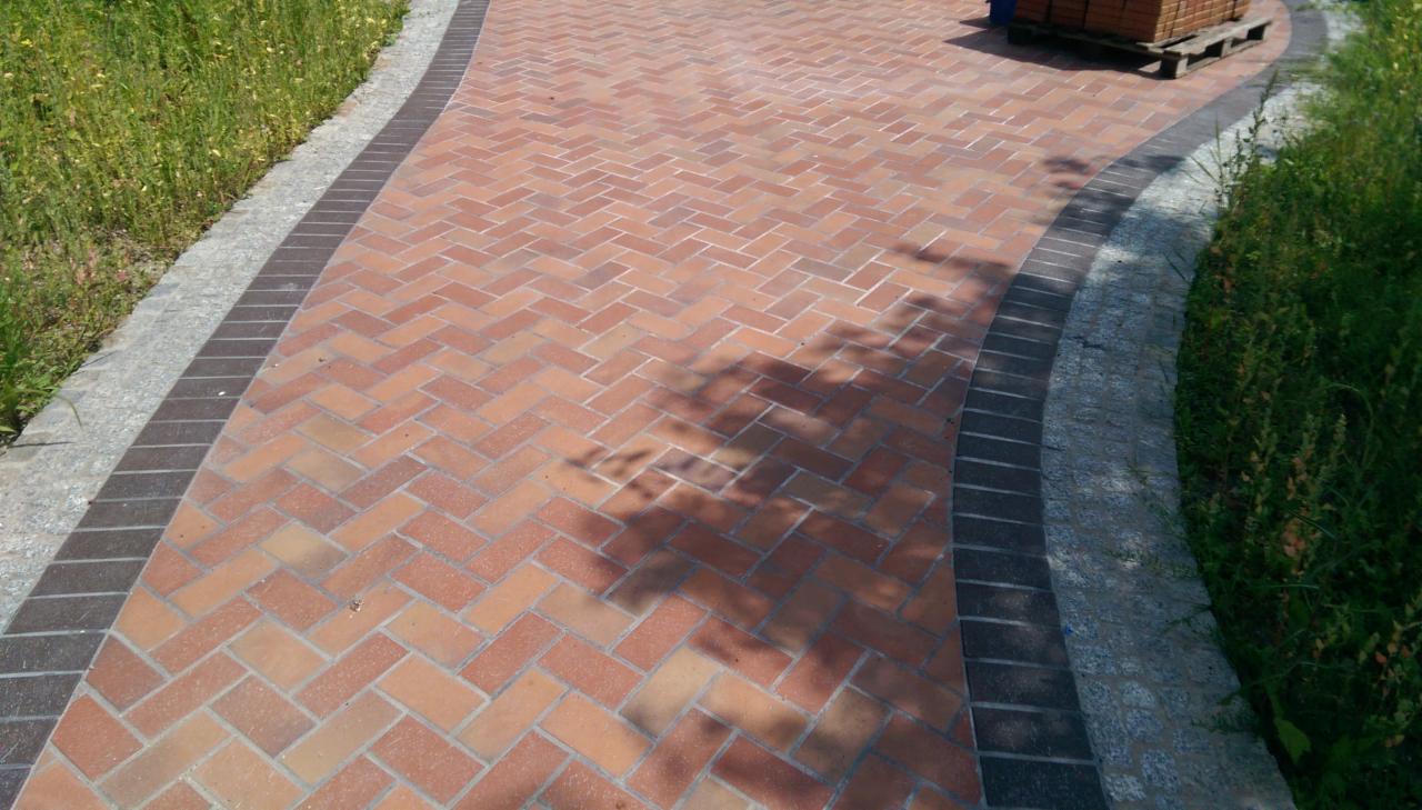 Ułożona z ceglastych kostek ścieżka w jodełkę, obramowana ciemniejszymi kostkami i szarym kamieniem, z paletą kostek w tle i cieniem drzewa na powierzchni.