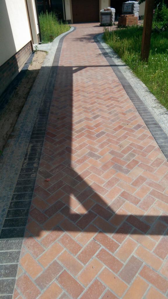 Nowo ułożona ścieżka z czerwonej kostki brukowej w jodełkę, obrzeża z ciemnoszarej kostki, cień drzewa na powierzchni, trawa i budynek w tle.