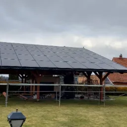 Montaż paneli słonecznych na drewnianej konstrukcji w ogrodzie, widoczne rusztowania i domy w tle.