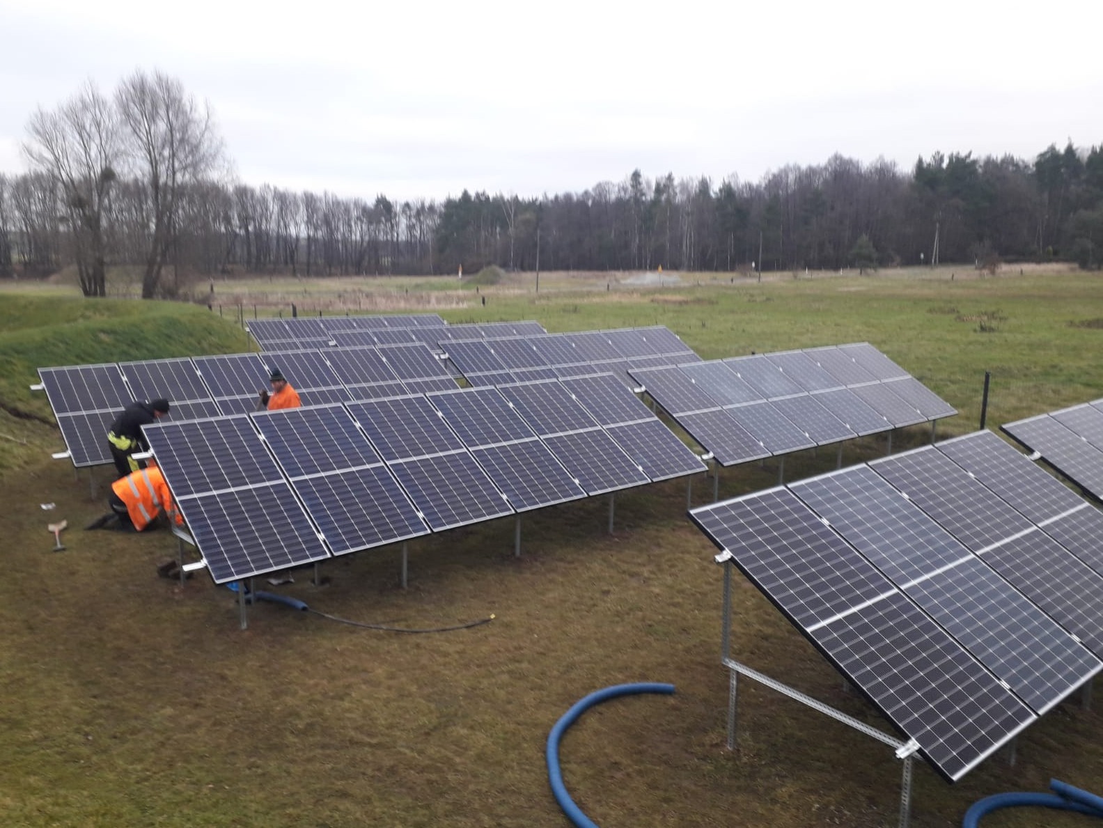Montaż paneli fotowoltaicznych na łące z widocznymi pracownikami w odblaskowych kamizelkach, niebieski wąż do podłączeń i metalowe konstrukcje wsporcze.