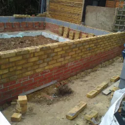 Budowa fundamentów z czerwonej i żółtej cegły, widoczne bloczki betonowe i rozkopana ziemia wewnątrz, etap murowania ścian.