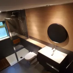 Nowoczesna łazienka z kabiną prysznicową typu walk-in, drewnianym siedziskiem, okrągłym lustrem i umywalką nablatową na szafce w odcieniach szarości i drewna.