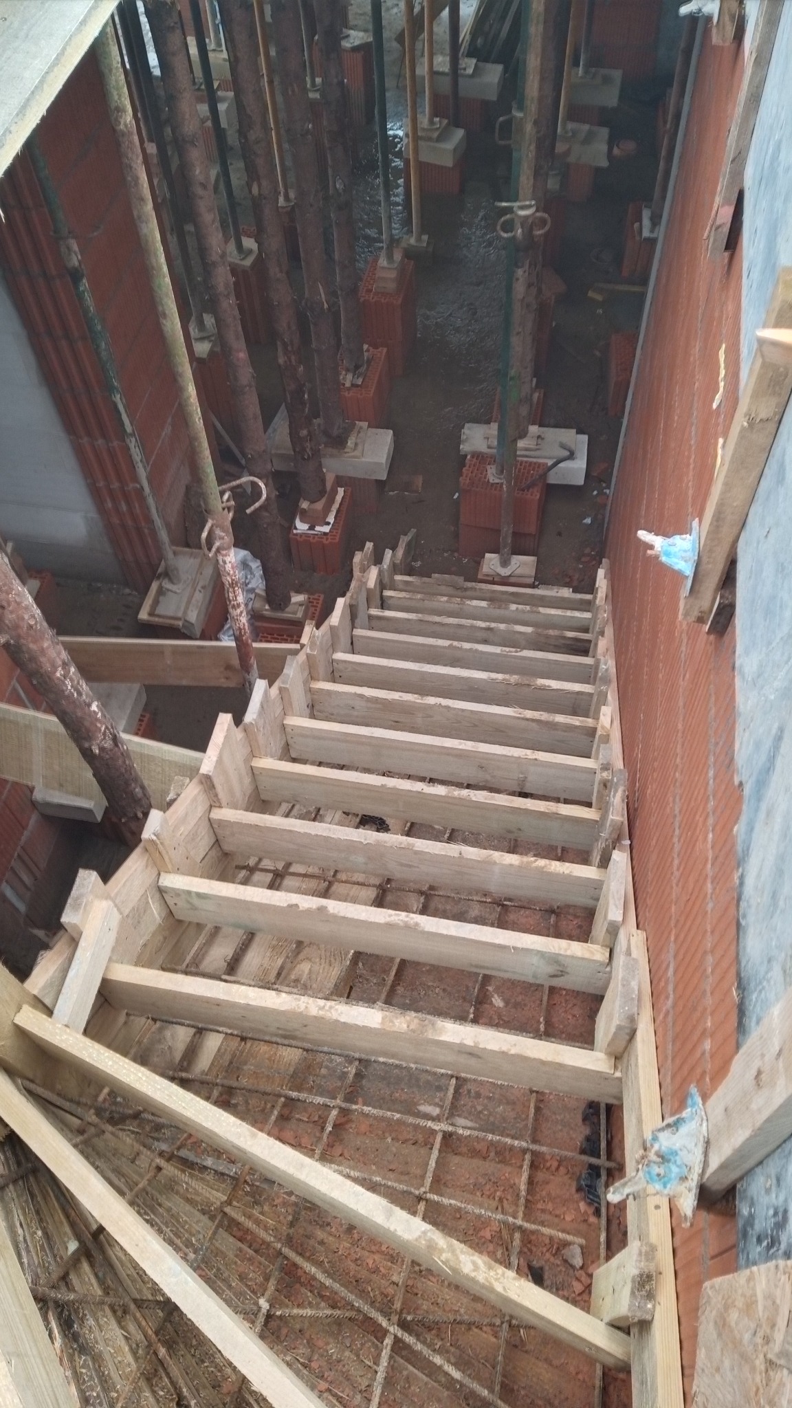 Szczegółowy widok z góry na drewniany szalunek schodów, zbrojenie w fundamencie i ceglane ściany w trakcie budowy domu murowanego.