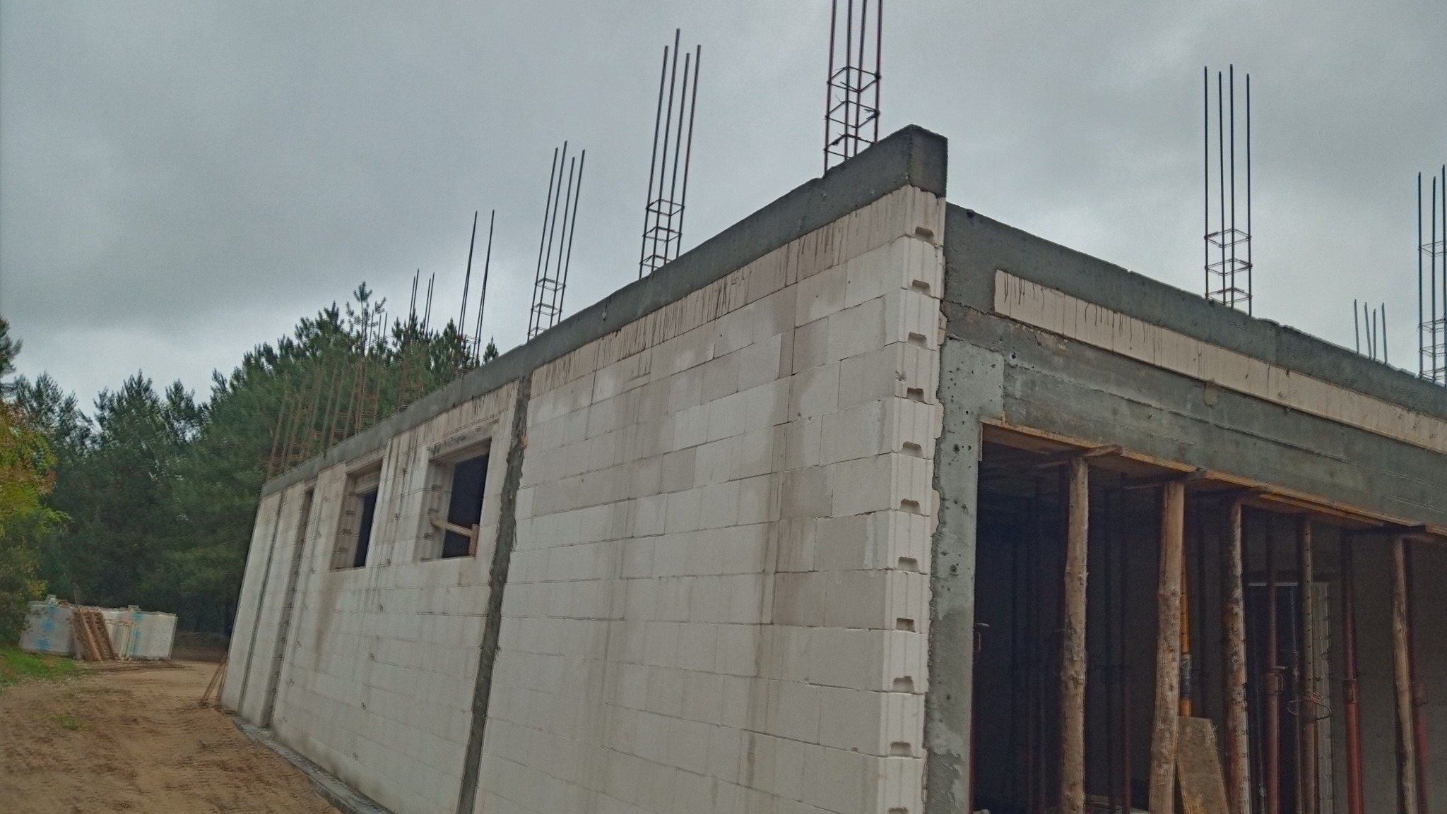 Surowy stan budynku z betonu komórkowego w trakcie budowy, widoczne wystające pręty zbrojeniowe i drewniane szalunki przy otworach, tło stanowią drzewa i pochmurne niebo.