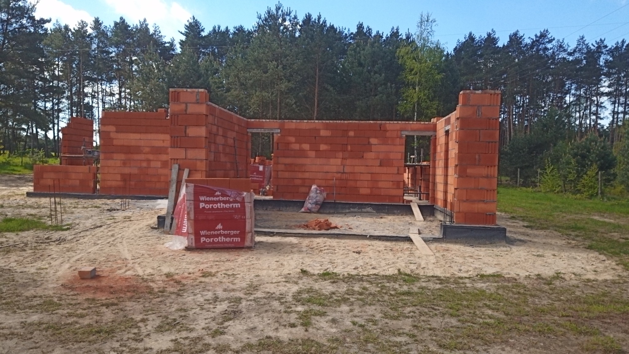 Wznoszone ściany domu z czerwonej cegły Porotherm, widoczne zbrojenia fundamentów, w tle las iglasty pod błękitnym niebem.