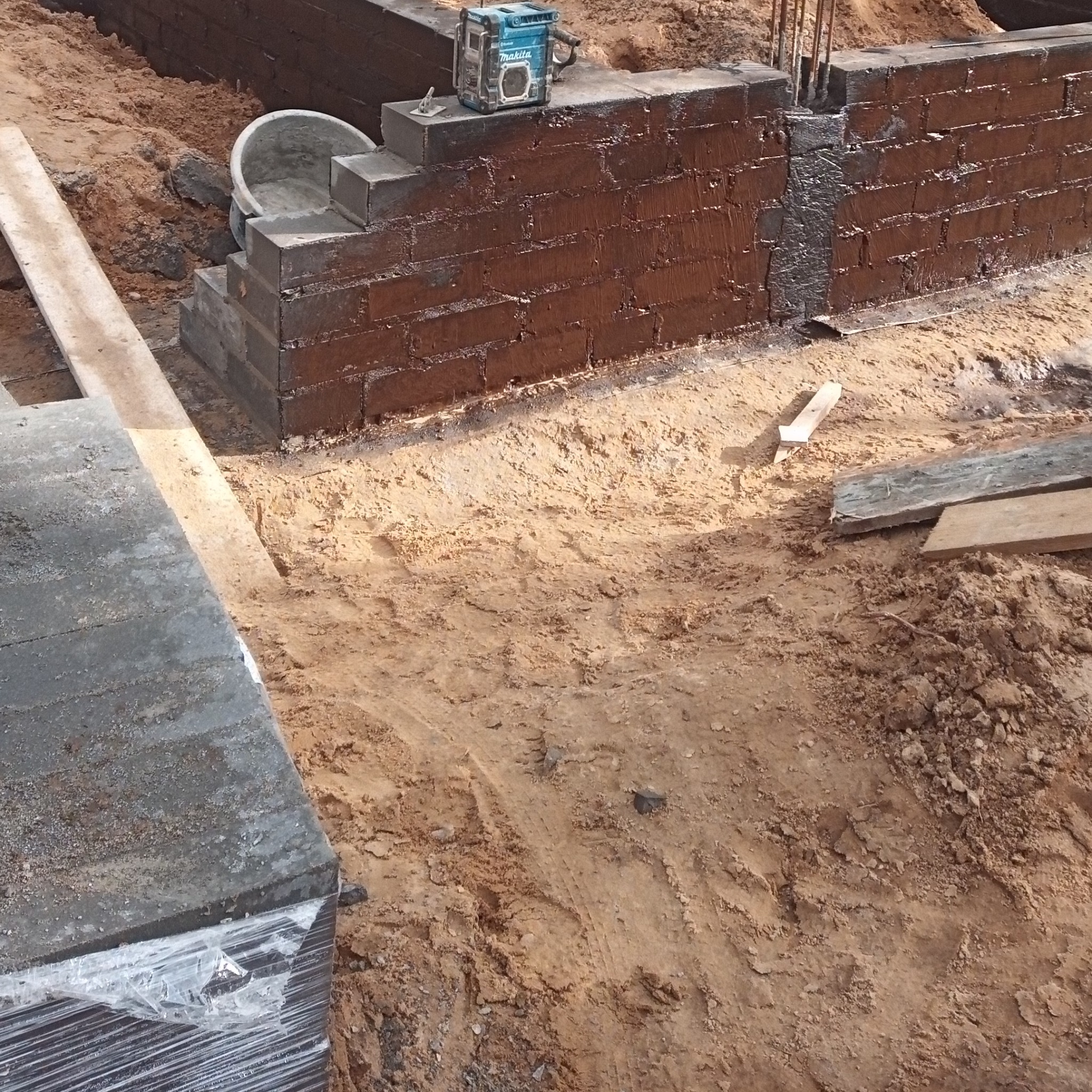 Wznoszenie fundamentów z bloczków betonowych, zabezpieczonych ciemną izolacją, z widocznym poziomnicą laserową Makita na górze i wiaderkiem z zaprawą obok, na placu budowy z piaszczystym podłożem.