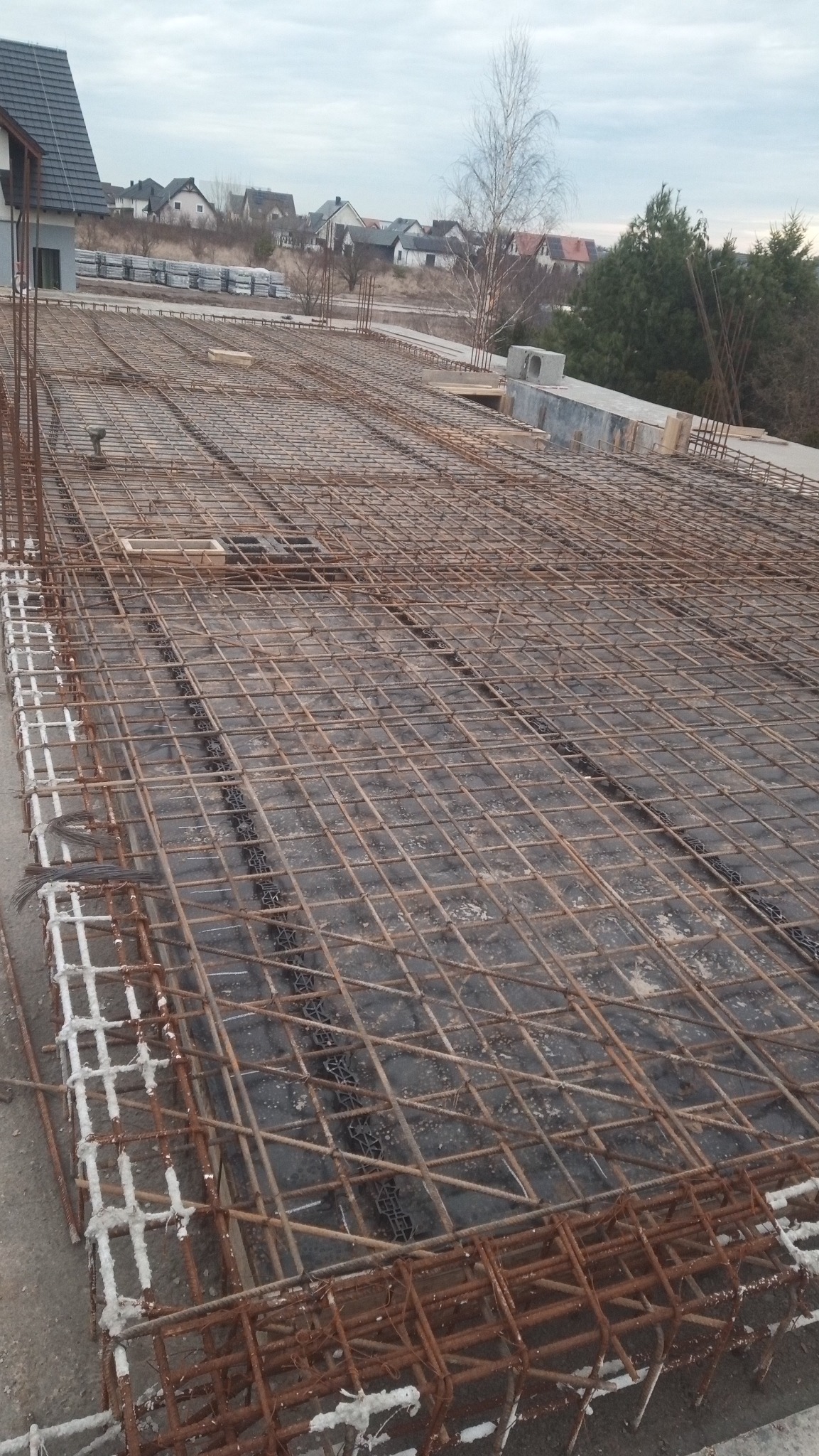 Zbrojenie płyty fundamentowej domu jednorodzinnego z widocznymi dystansami z tworzywa sztucznego, na tle zabudowy jednorodzinnej w pochmurny dzień.