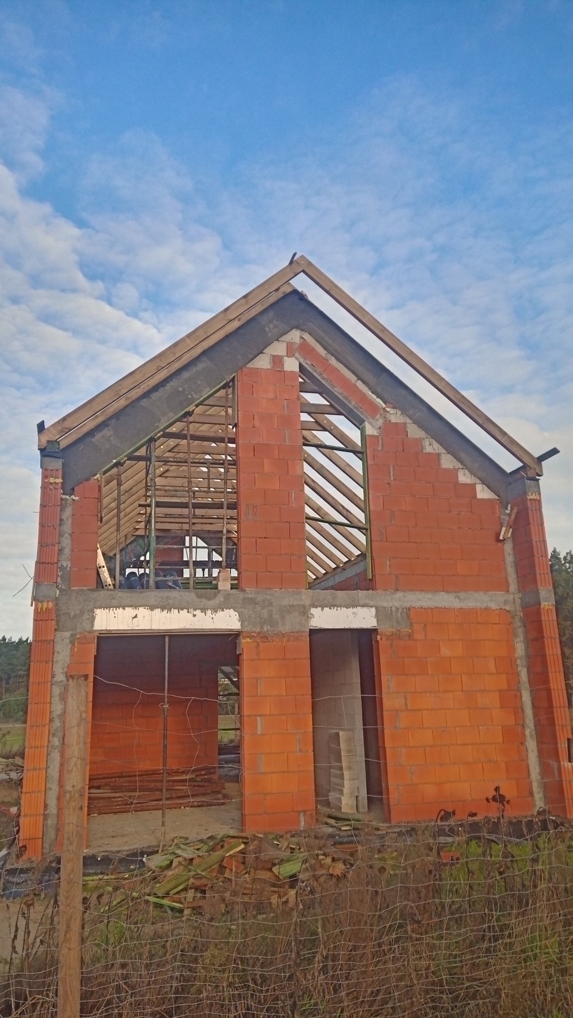 Dwukondygnacyjny budynek w stanie surowym otwartym z czerwonej cegły, widoczne drewniane elementy konstrukcji dachu i stropu, siatka ogrodzeniowa w pierwszym planie, błękitne niebo w tle.
