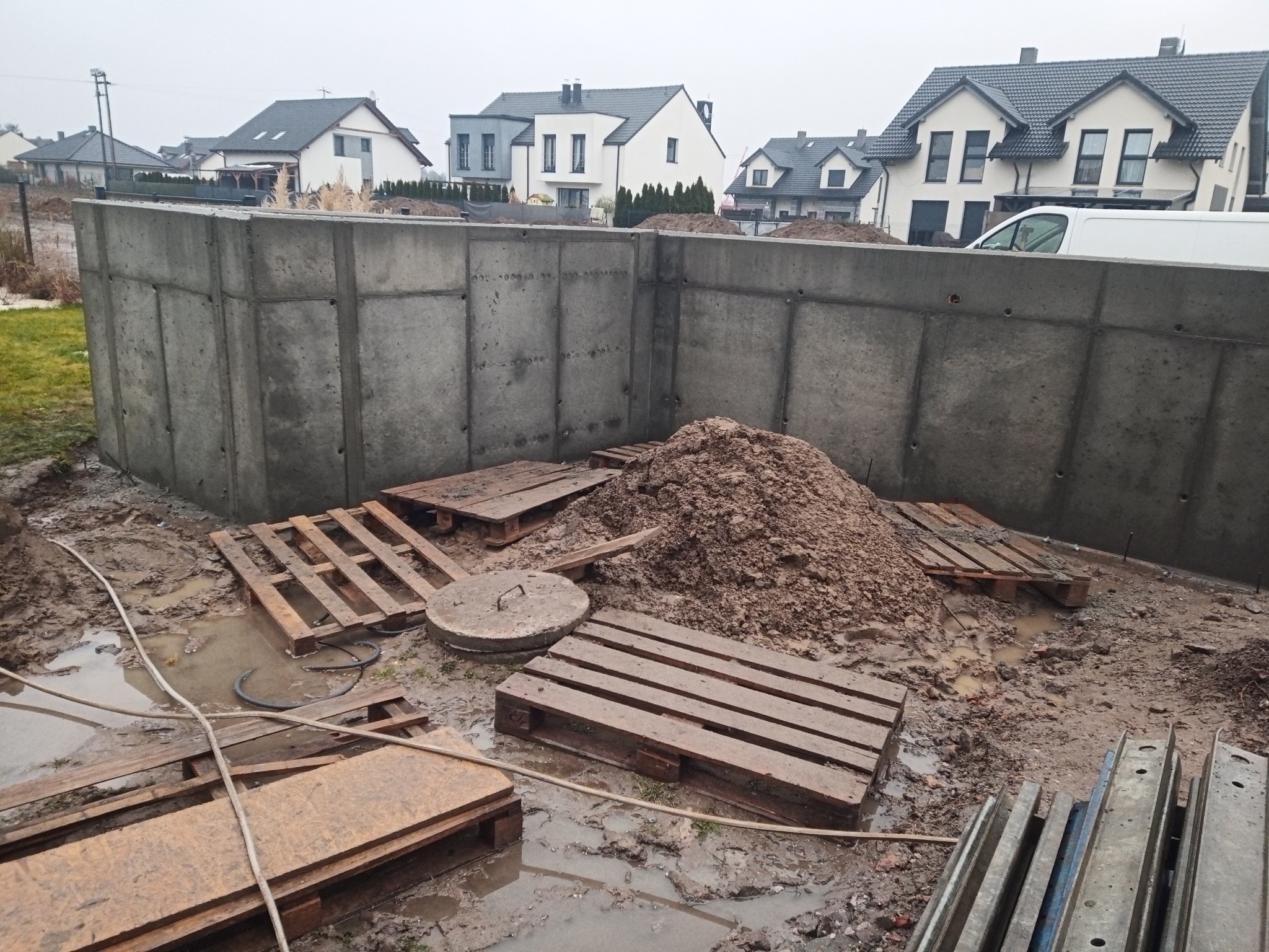 Wylane fundamenty segmentowe z betonu, widoczne szalunki, palety i ziemia. W tle zabudowa jednorodzinna w pochmurny dzień.
