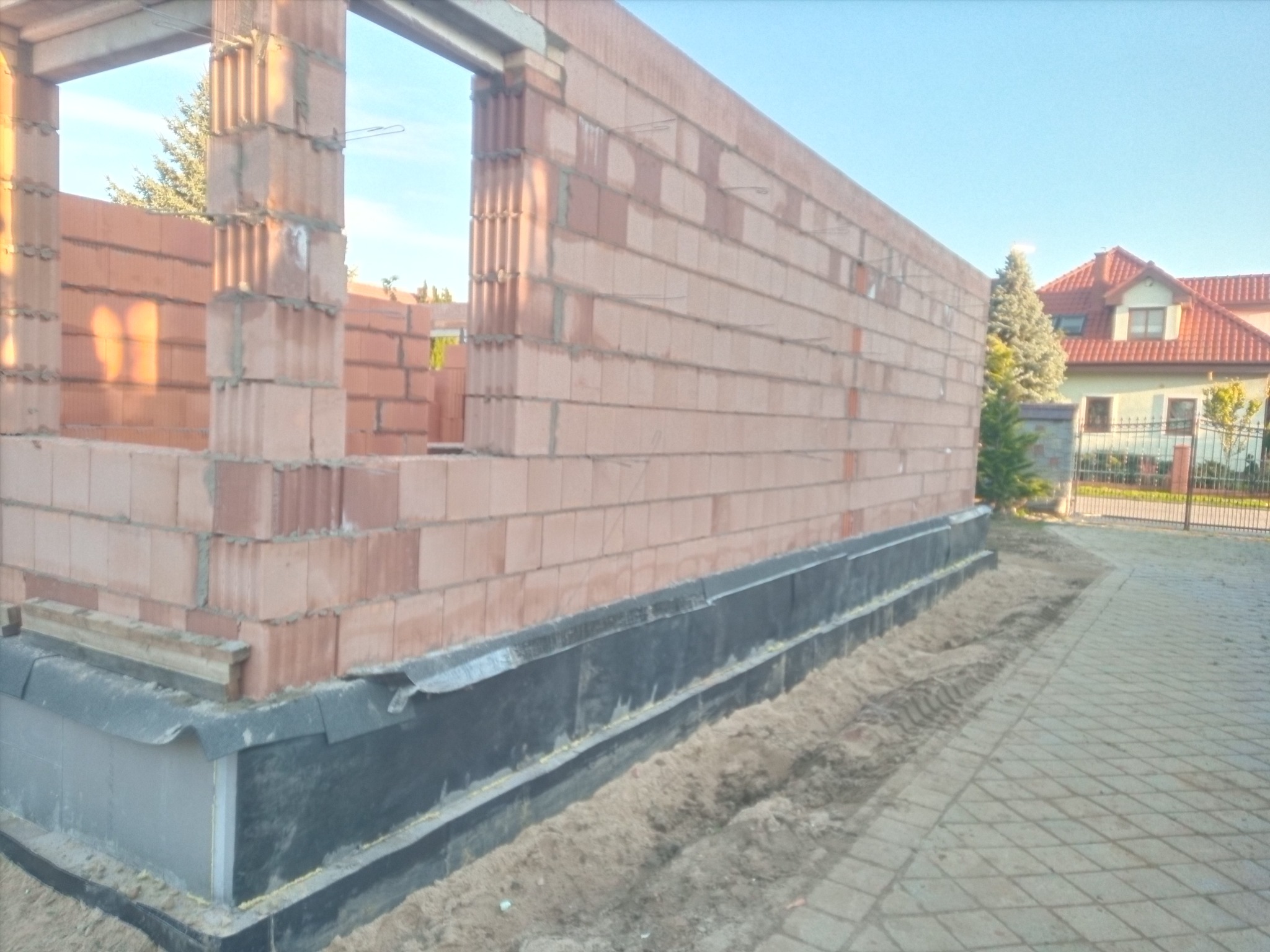 Wznoszone ściany z czerwonej cegły porotherm na fundamencie z izolacją przeciwwilgociową. Widok na budynek w tle i kostkę brukową.