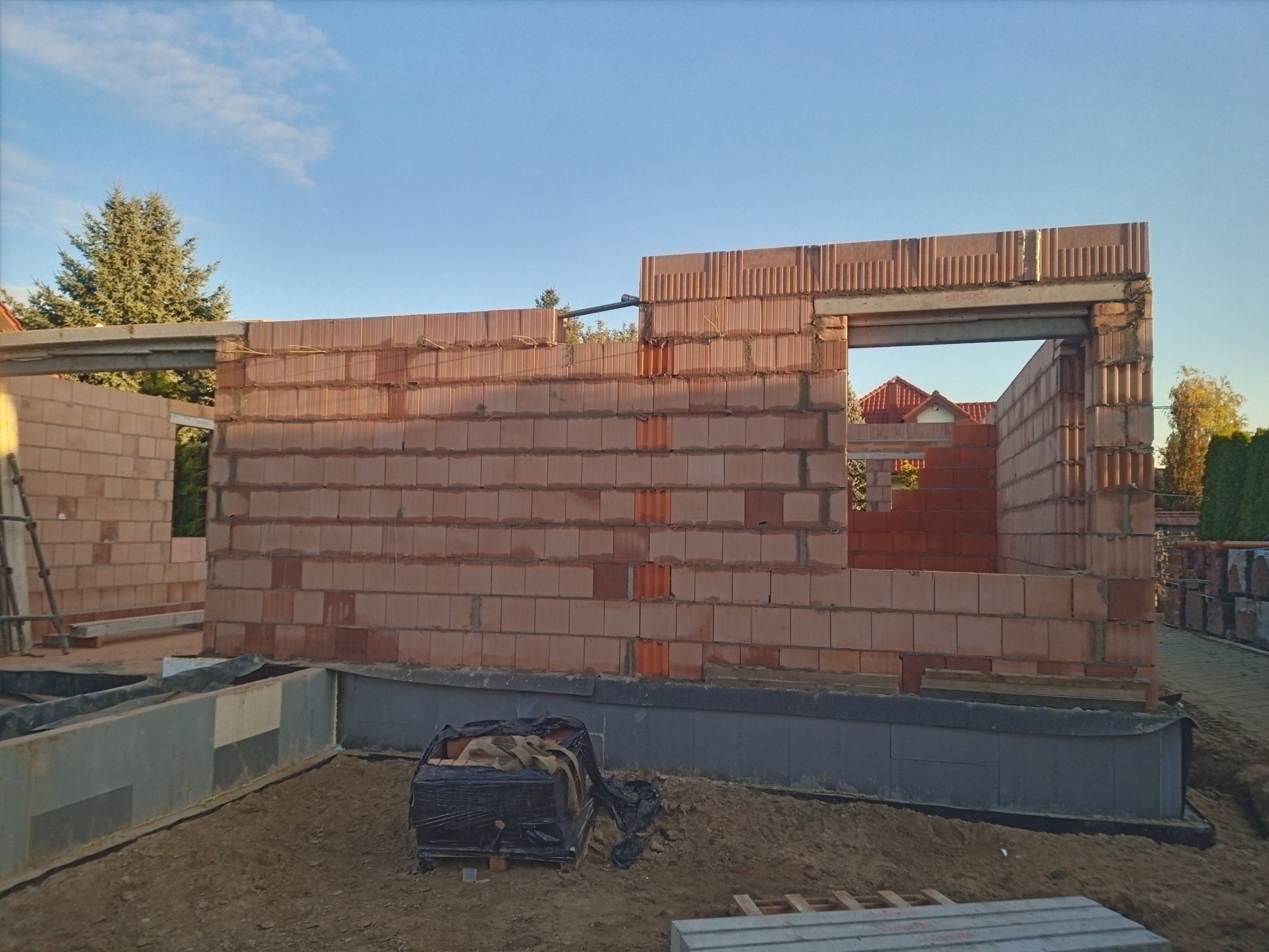 Wznoszenie ścian domu z czerwonej cegły ceramicznej. Widoczne fundamenty, otwory okienne i nadproża. Częściowo zrealizowana budowa domu jednorodzinnego w słoneczny dzień.