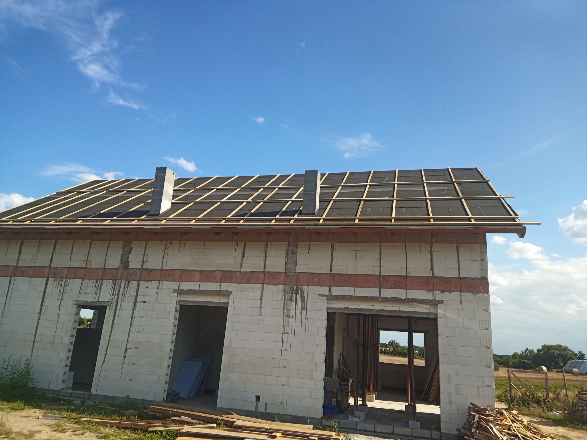 Niedokończony dom z białego pustaka, widoczne drewniane elementy konstrukcji dachu i komin. W tle błękitne niebo z chmurami.