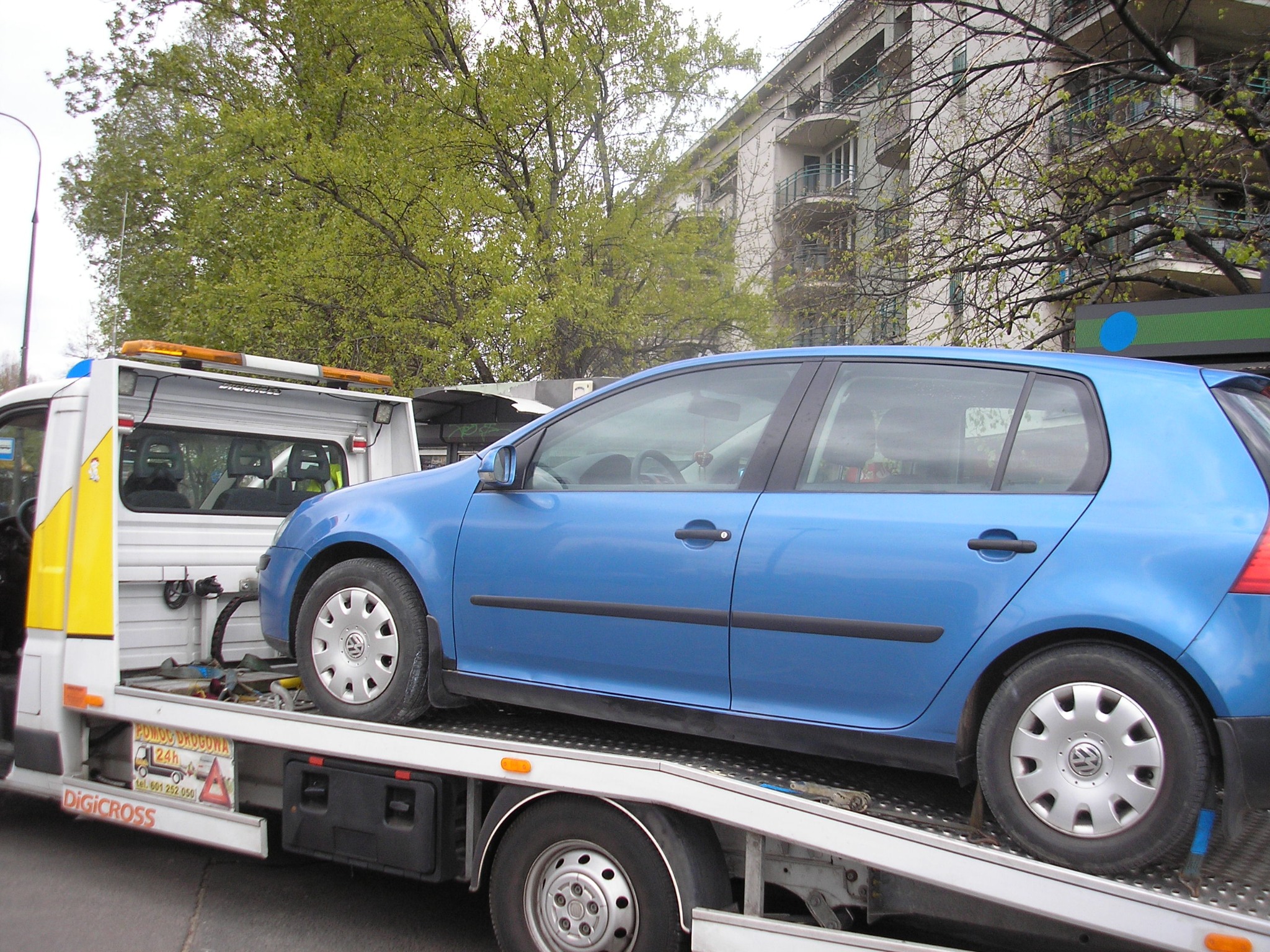 Niebieski Volkswagen Golf załadowany na lawetę w Warszawie, z widocznym budynkiem mieszkalnym w tle.
