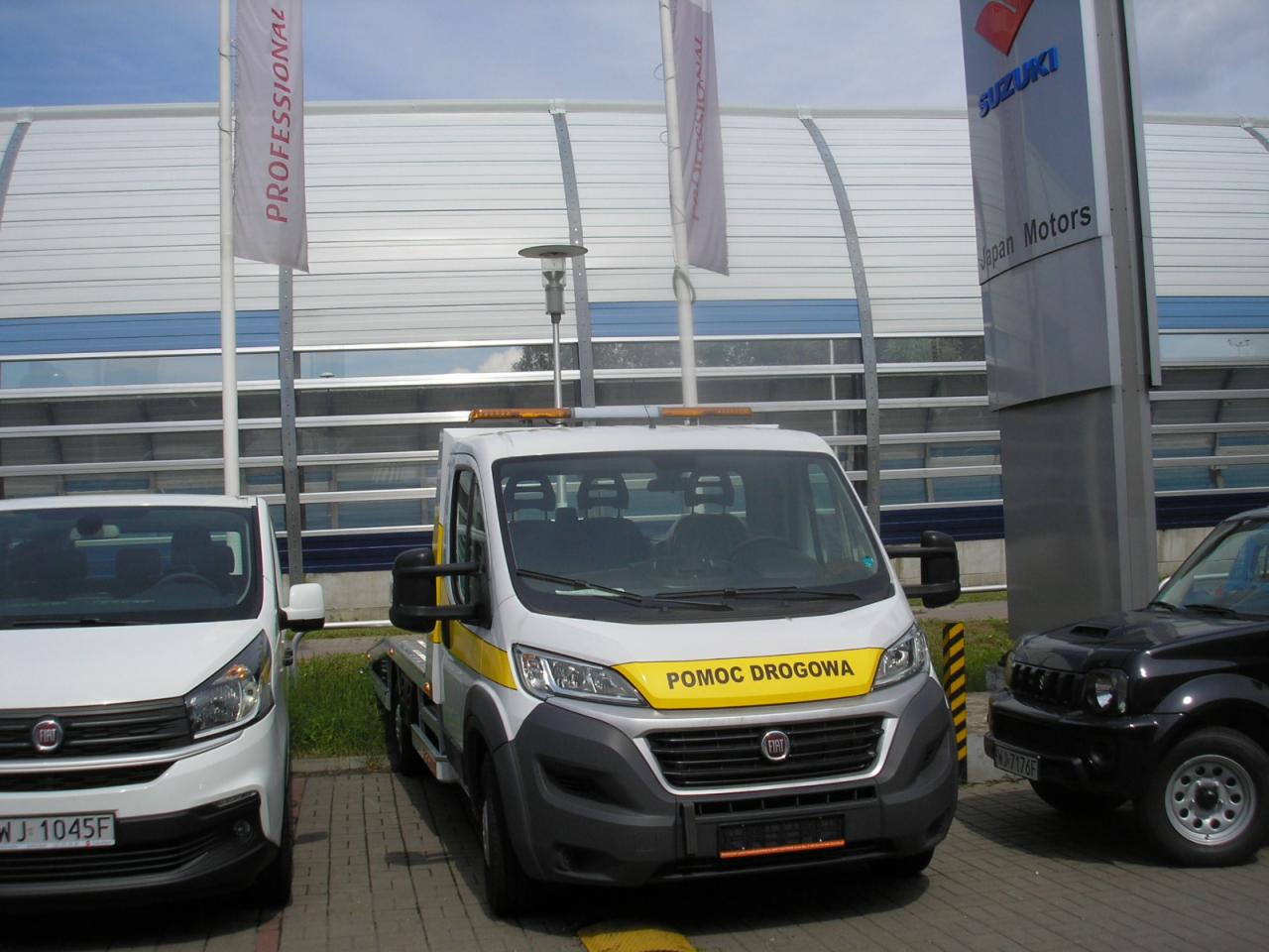 Nowa, biała laweta z żółtym pasem i napisem POMOC DROGOWA na tle salonu samochodowego Suzuki, zaparkowana obok białego Fiata i czarnego Suzuki Jimny.