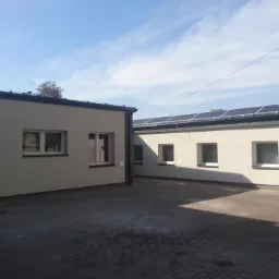 Dwa budynki biurowe z panelami słonecznymi na dachu, świeżo po remoncie, z widoczną nową elewacją w odcieniach beżu i szarości, oraz kostką brukową na dziedzińcu.