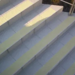 Schody wyłożone szarymi płytkami z żółtymi pasami antypoślizgowymi, oświetlone promieniami słońca, z metalową barierką w tle.