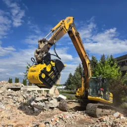 Żółta koparka z frezem hydraulicznym rozbiera gruzowisko na tle błękitnego nieba i budynku. Widoczne ślady rozbiórki i operator w kabinie maszyny.