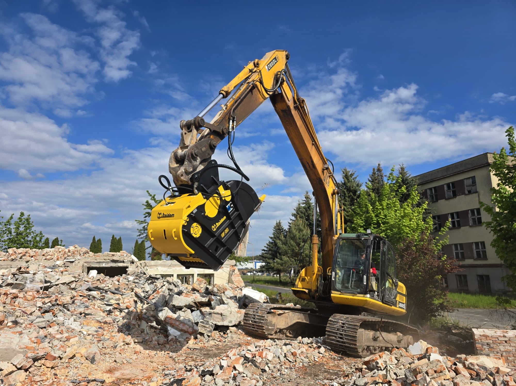 Żółta koparka z frezem hydraulicznym rozbiera gruzowisko na tle błękitnego nieba i budynku. Widoczne ślady rozbiórki i operator w kabinie maszyny.