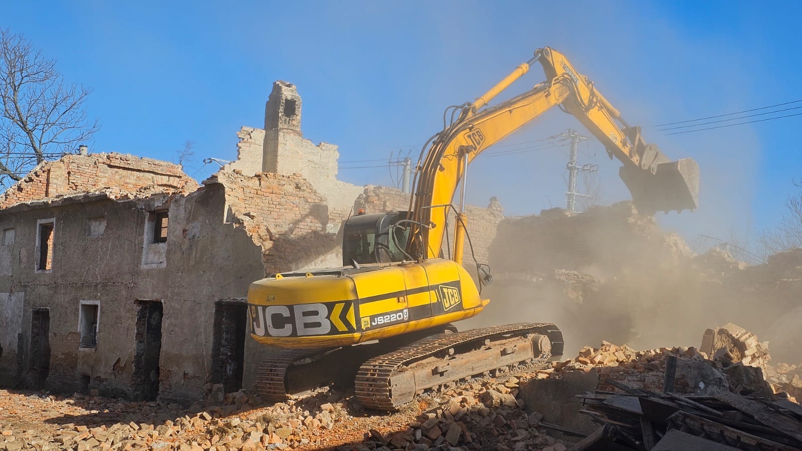 Żółta koparka JCB JS220 burzy ceglany budynek, unosząc tumany kurzu. Częściowo zburzona konstrukcja z widocznymi oknami i kominem na tle błękitnego nieba.