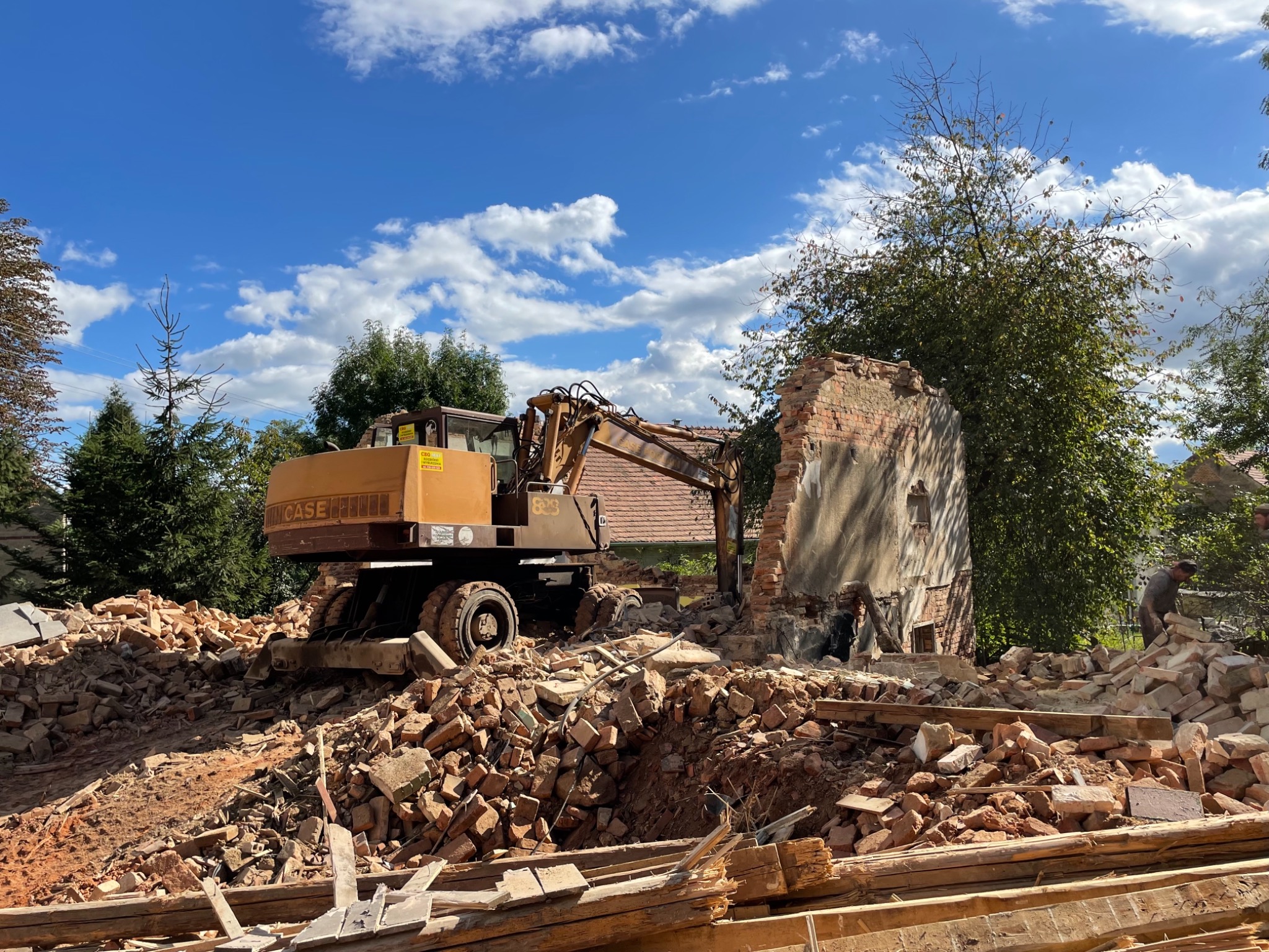 Żółta koparka CASE na tle częściowo zburzonej ceglanej ściany i sterty gruzu, z widocznym niebieskim niebem i drzewami w tle.