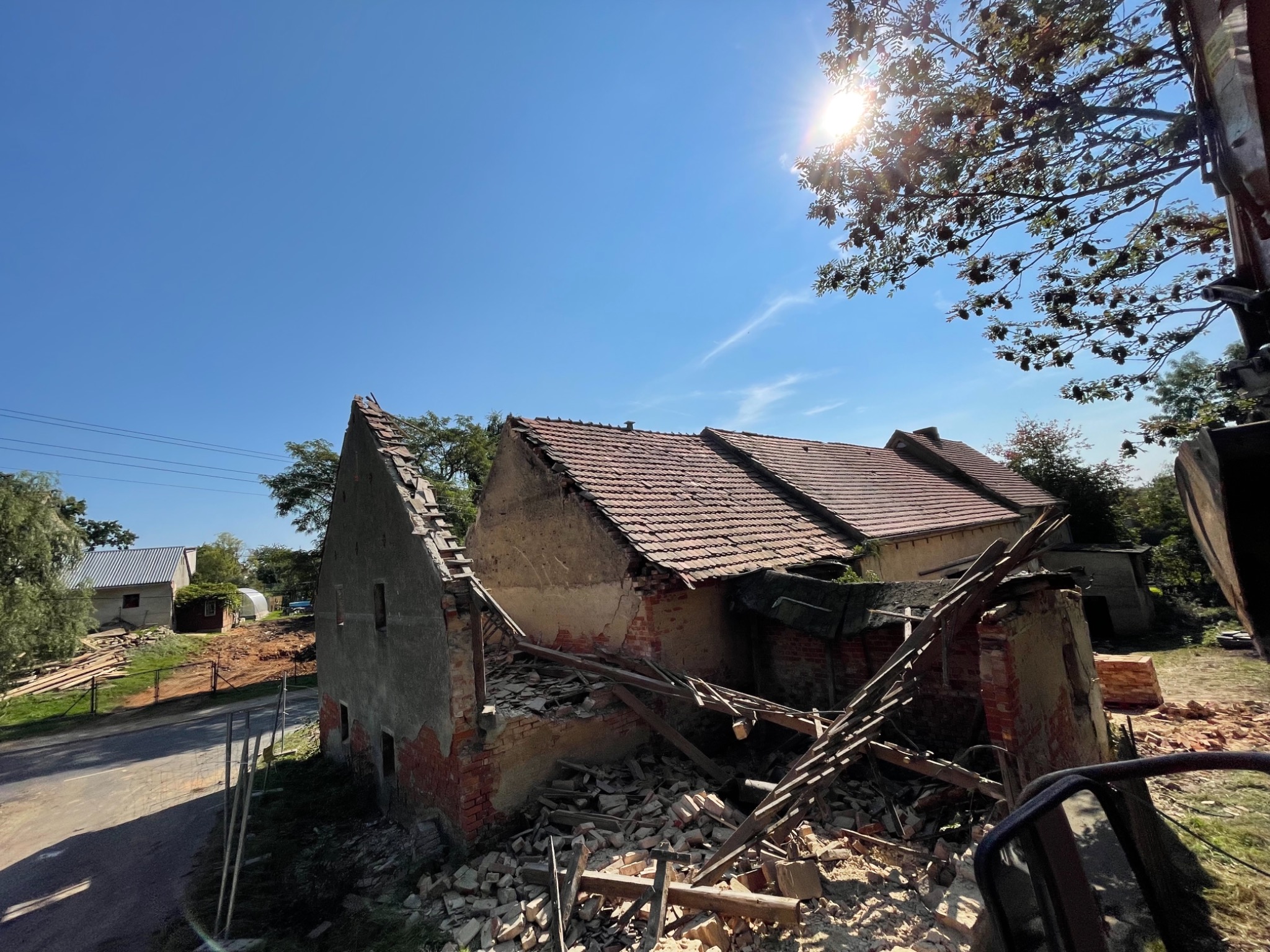 Częściowo zburzony ceglany budynek z czerwoną dachówką, z widocznymi gruzami i elementami konstrukcji, sfotografowany w słoneczny dzień z perspektywy maszyny budowlanej.