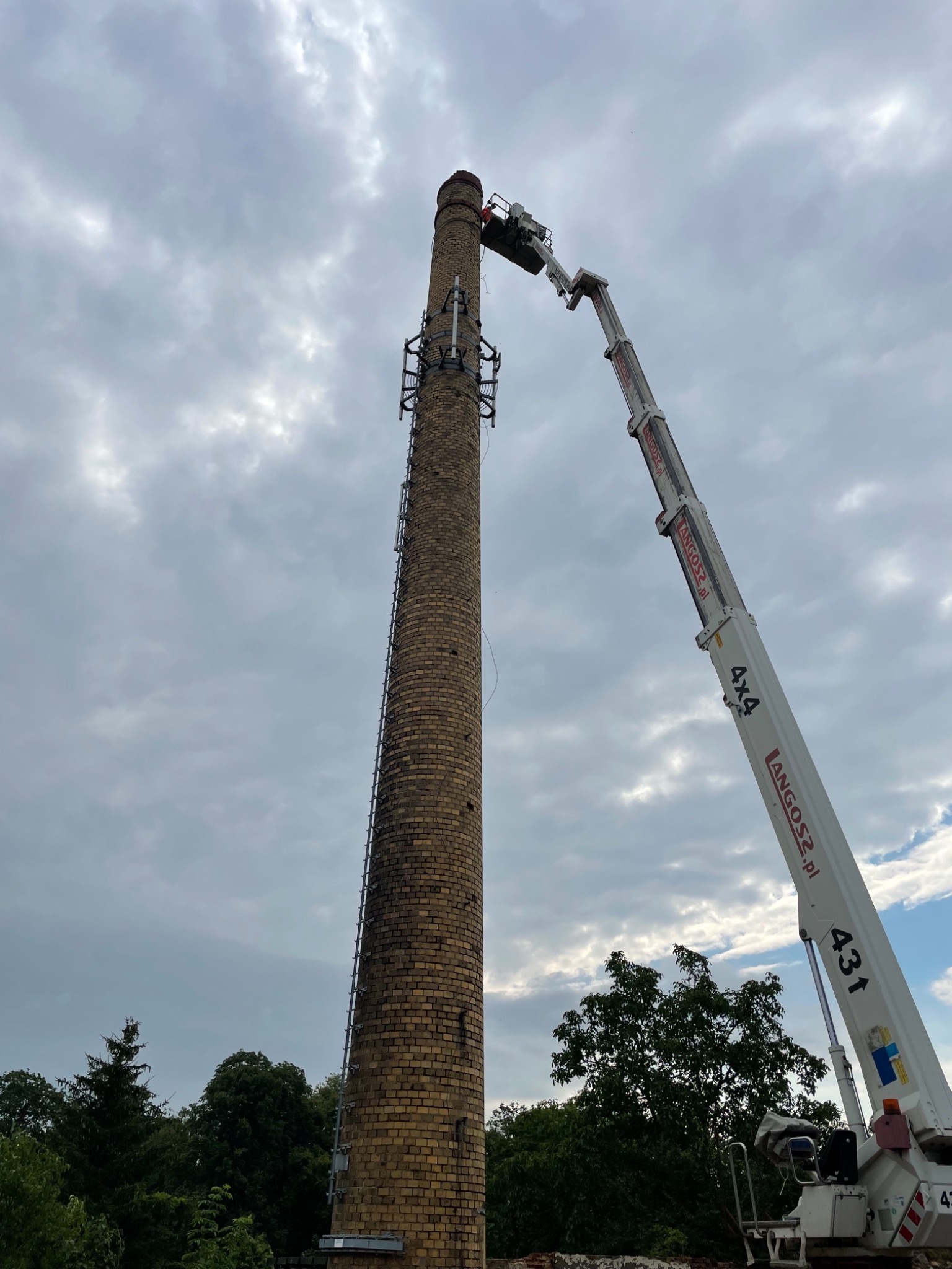 Wysoki ceglany komin przemysłowy z zamontowaną drabiną, obsługiwany przez biały podnośnik koszowy z wysięgnikiem, na tle pochmurnego nieba.