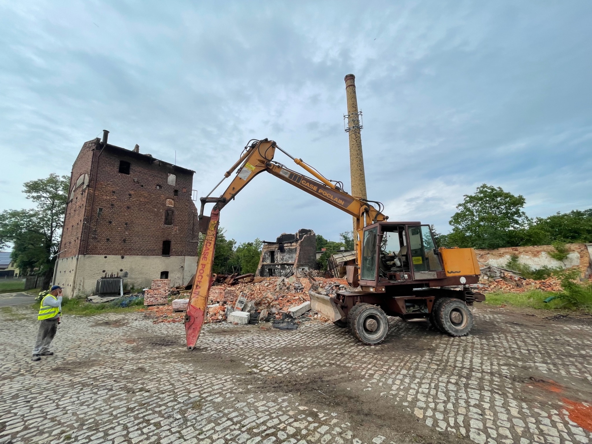 Żółta koparka Case Poclain na tle częściowo zburzonego, ceglanego budynku z kominem, pracownik w żółtej kamizelce obserwuje postęp prac.