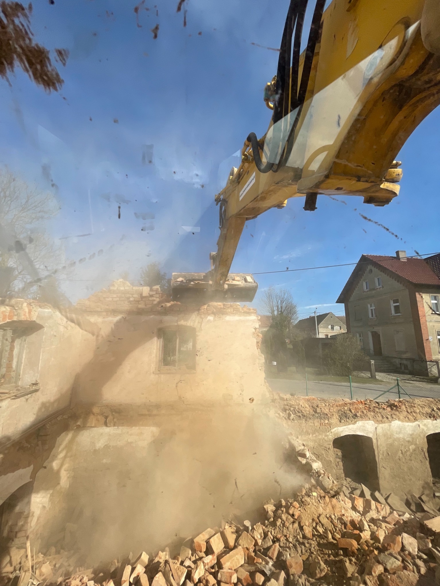 Żółta koparka burzy ceglany budynek, widok z perspektywy operatora, pył i gruz unoszą się w powietrzu, w tle zabudowa mieszkalna.
