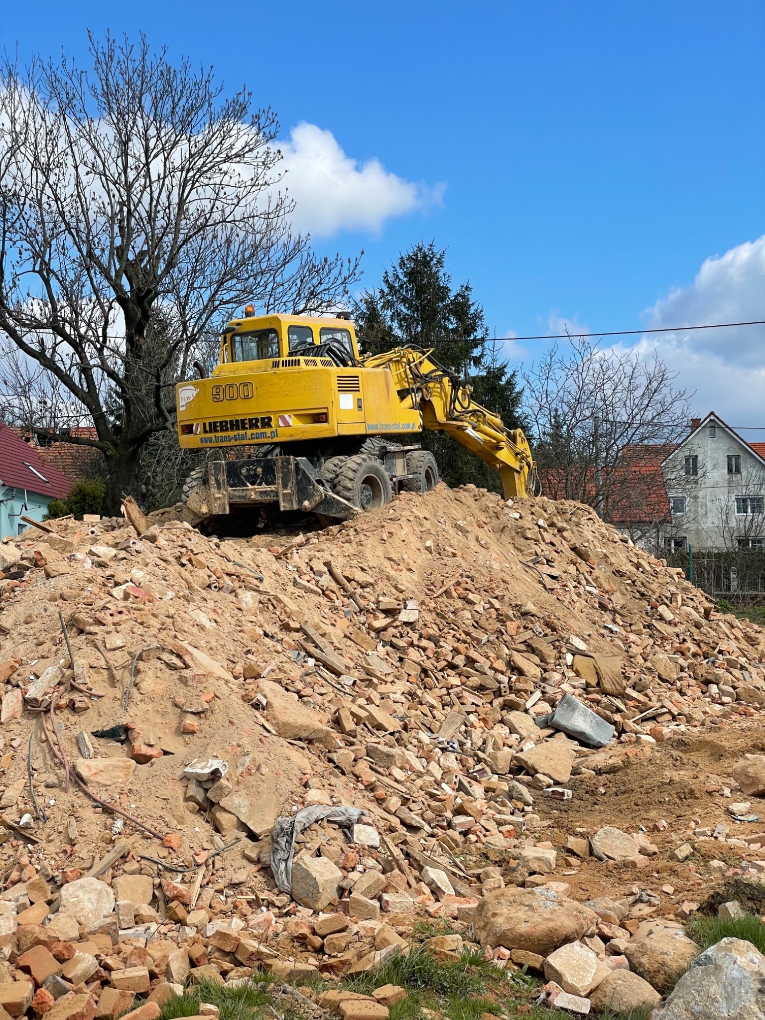 Żółta koparka Liebherr 900 stoi na stercie gruzu i cegieł po rozbiórce, na tle błękitnego nieba z chmurami i drzew bez liści.