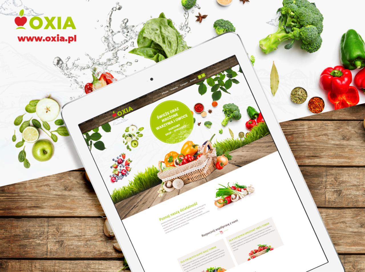 Tablet wyświetla stronę internetową firmy Oxia z owocami i warzywami, otoczony świeżymi produktami spożywczymi na białym tle z drewnianym akcentem.