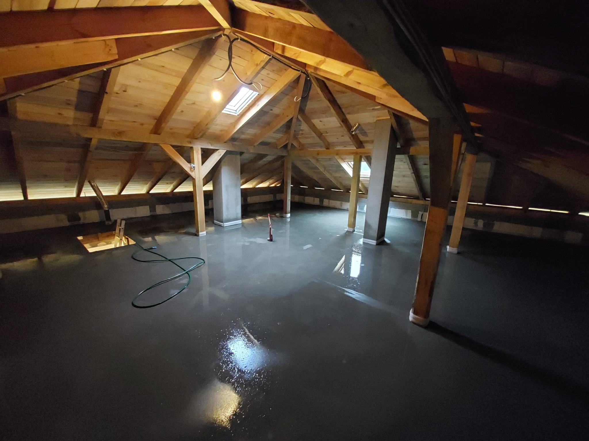 Wylewka samopoziomująca na poddaszu z drewnianą konstrukcją dachu i oknem dachowym, widoczny wąż i narzędzie do rozprowadzania wylewki.