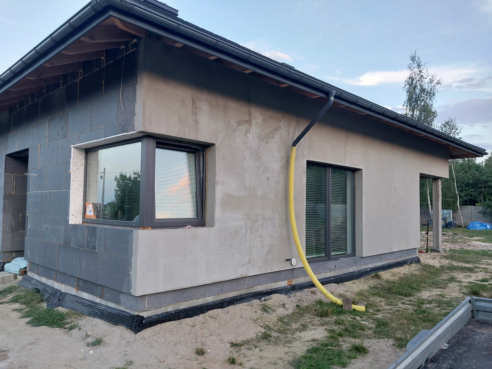 Surowy stan elewacji domu jednorodzinnego z widocznymi oknami z roletami, częściowo ocieplonego szarym styropianem, z żółtą rurą spustową i podsypką wokół fundamentów.
