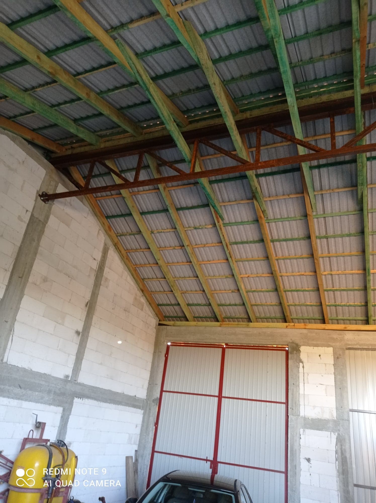 Wnętrze hali w trakcie budowy: widoczne białe ściany z betonu komórkowego, drewniana konstrukcja dachu z zielonymi impregnatami i szara blacha falista, czerwona brama wjazdowa, częściowo widoczny...