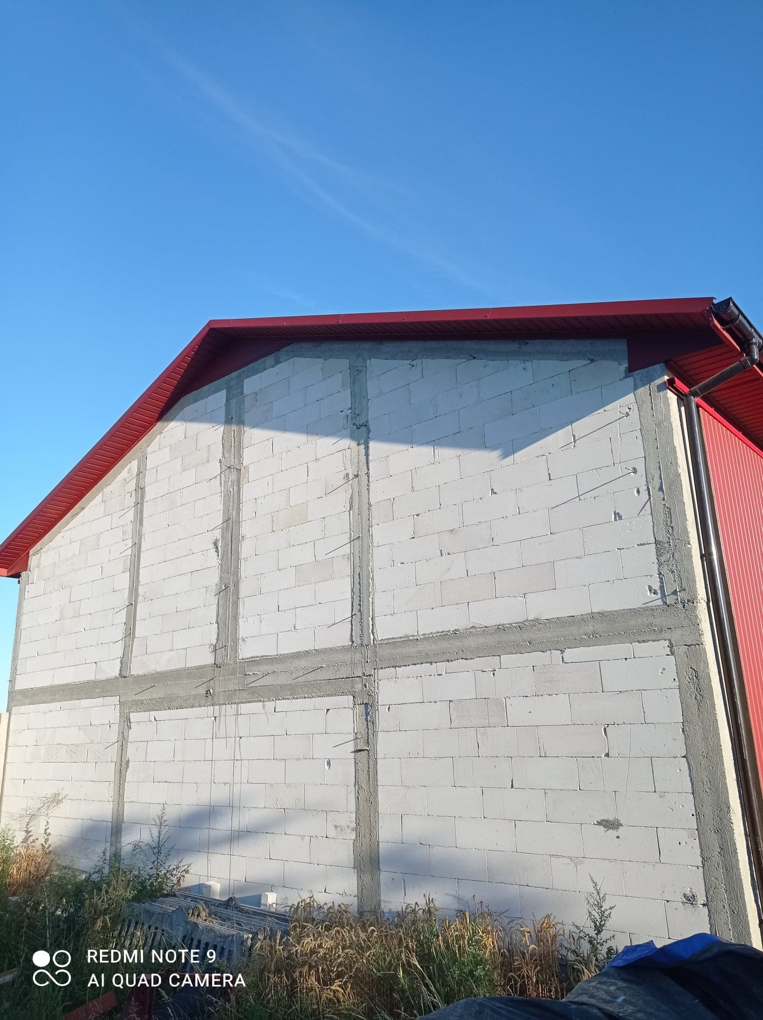 Surowy stan budynku z białych bloczków, z widoczną konstrukcją wzmacniającą i czerwonym dachem, na tle błękitnego nieba.