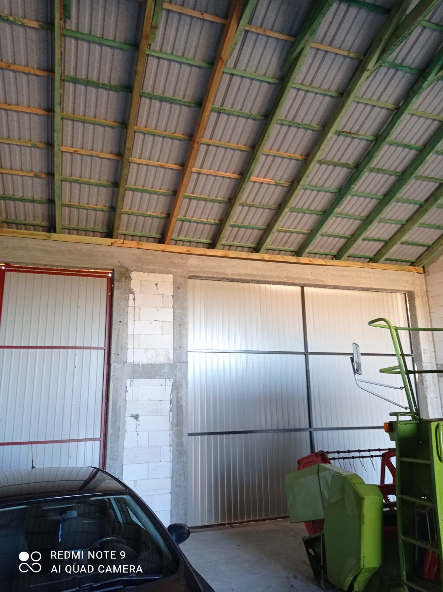 Wnętrze budynku gospodarczego w trakcie budowy: widoczne ściany z bloczków, metalowe wrota, konstrukcja dachu z blachy trapezowej i drewnianych belek, fragment samochodu i maszyny rolniczej.