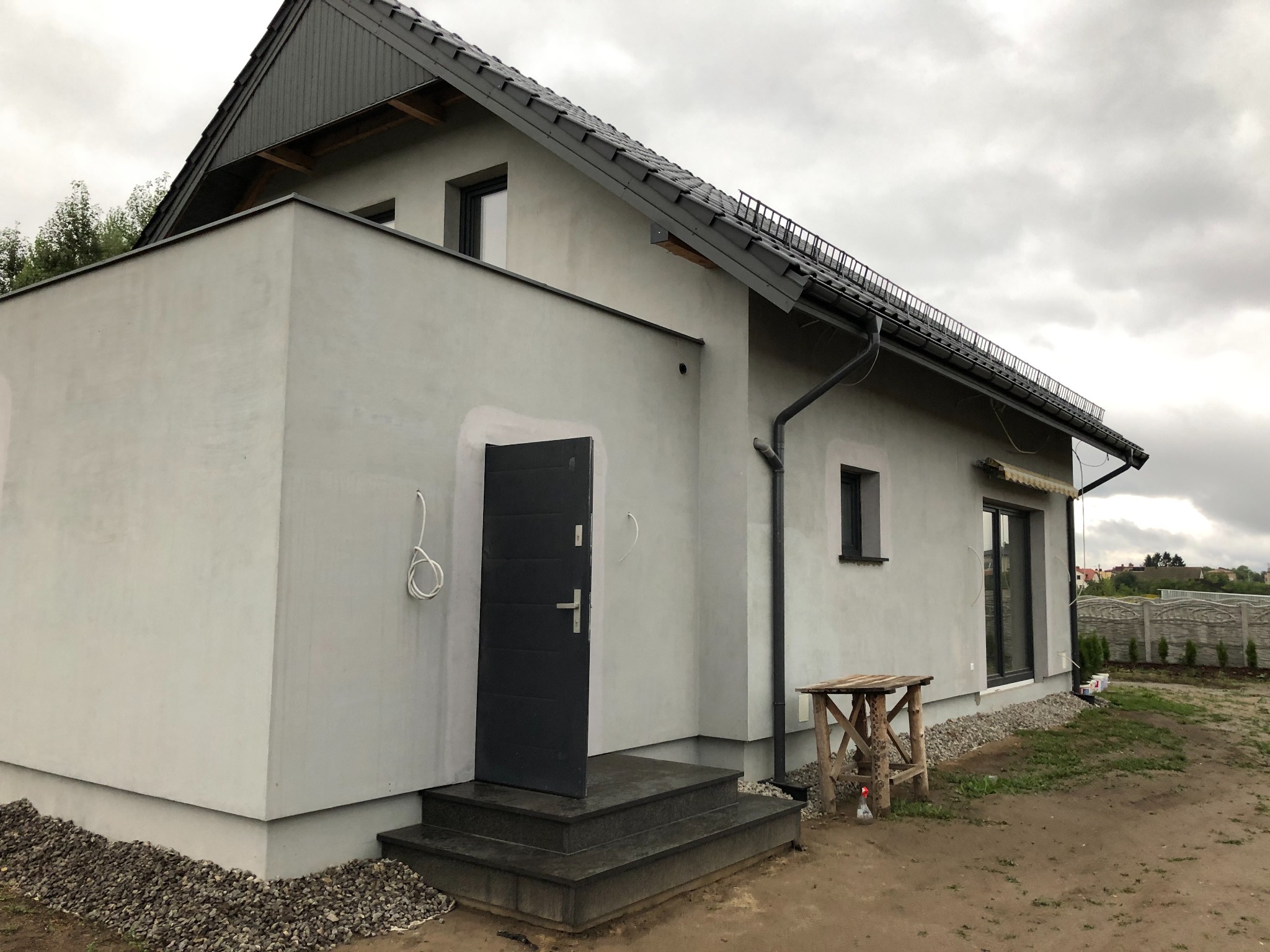 Nowoczesny dom jednorodzinny z szarą elewacją i ciemną stolarką okienną, widoczne schody wejściowe z ciemnego kamienia i drewniany stół roboczy na zewnątrz.