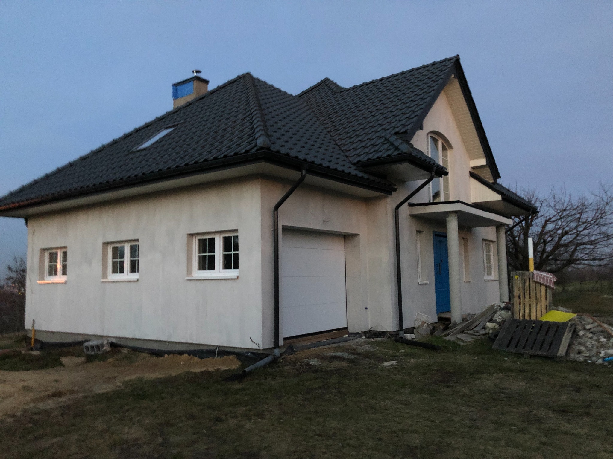 Niedokończony dom jednorodzinny z garażem, widoczne materiały budowlane, elewacja w trakcie prac, niebieskie drzwi wejściowe, szara dachówka.