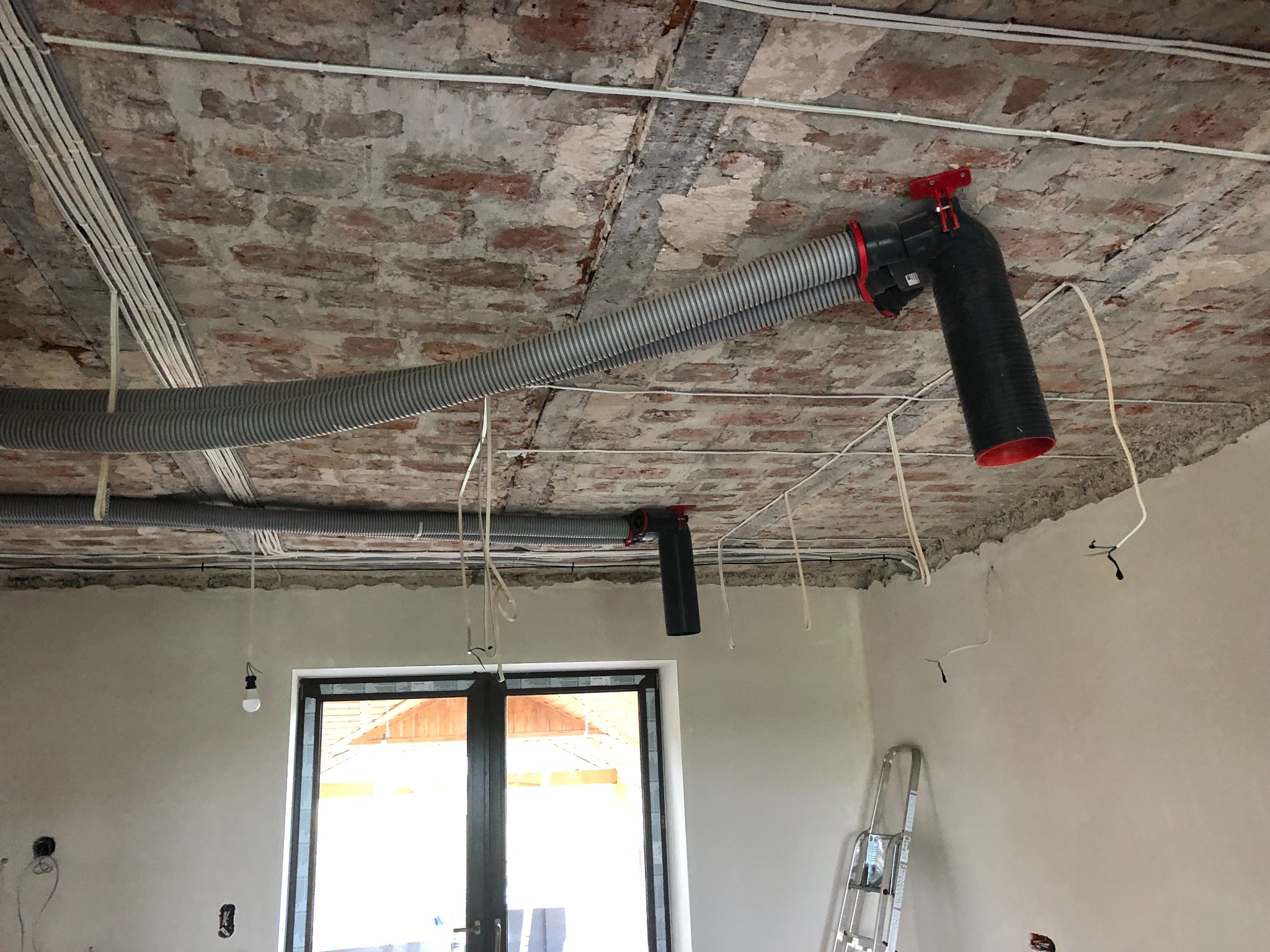 Montaż systemu wentylacji mechanicznej z odzyskiem ciepła: widoczne rury wentylacyjne pod sufitem z surowej cegły, czarne anemostaty z czerwonymi elementami, instalacja elektryczna w trakcie prac...