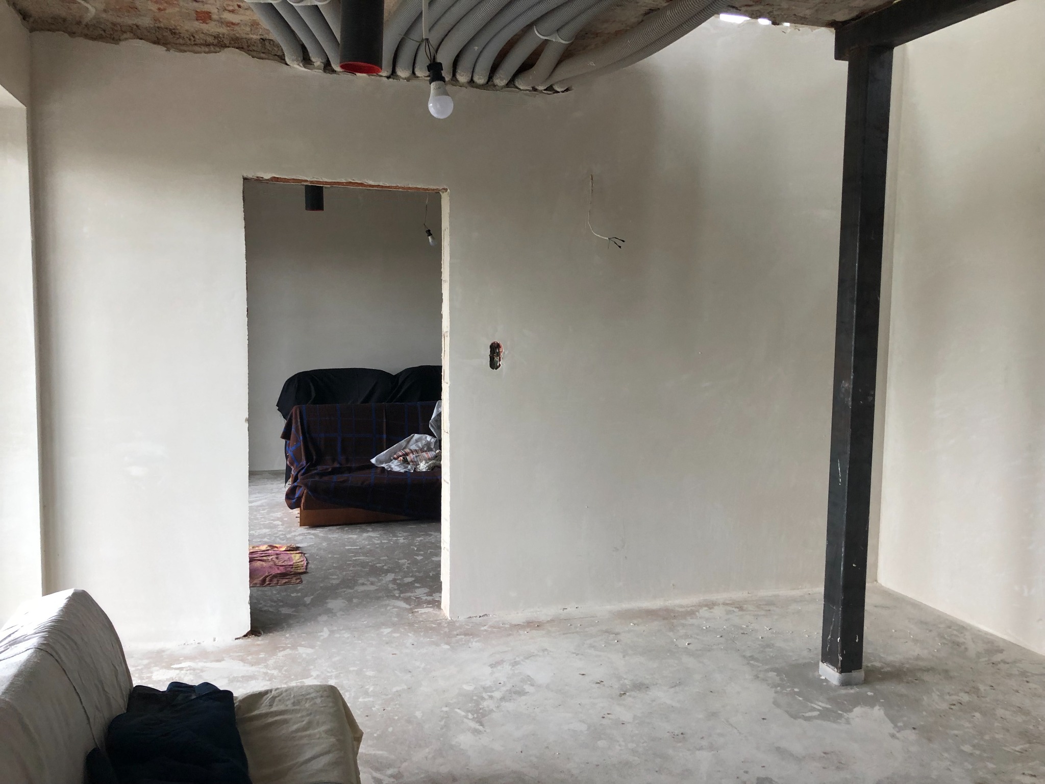 Wnętrze w trakcie remontu: świeżo otynkowane ściany, odsłonięty strop z instalacjami, surowa betonowa podłoga i stalowa kolumna w rogu pomieszczenia.