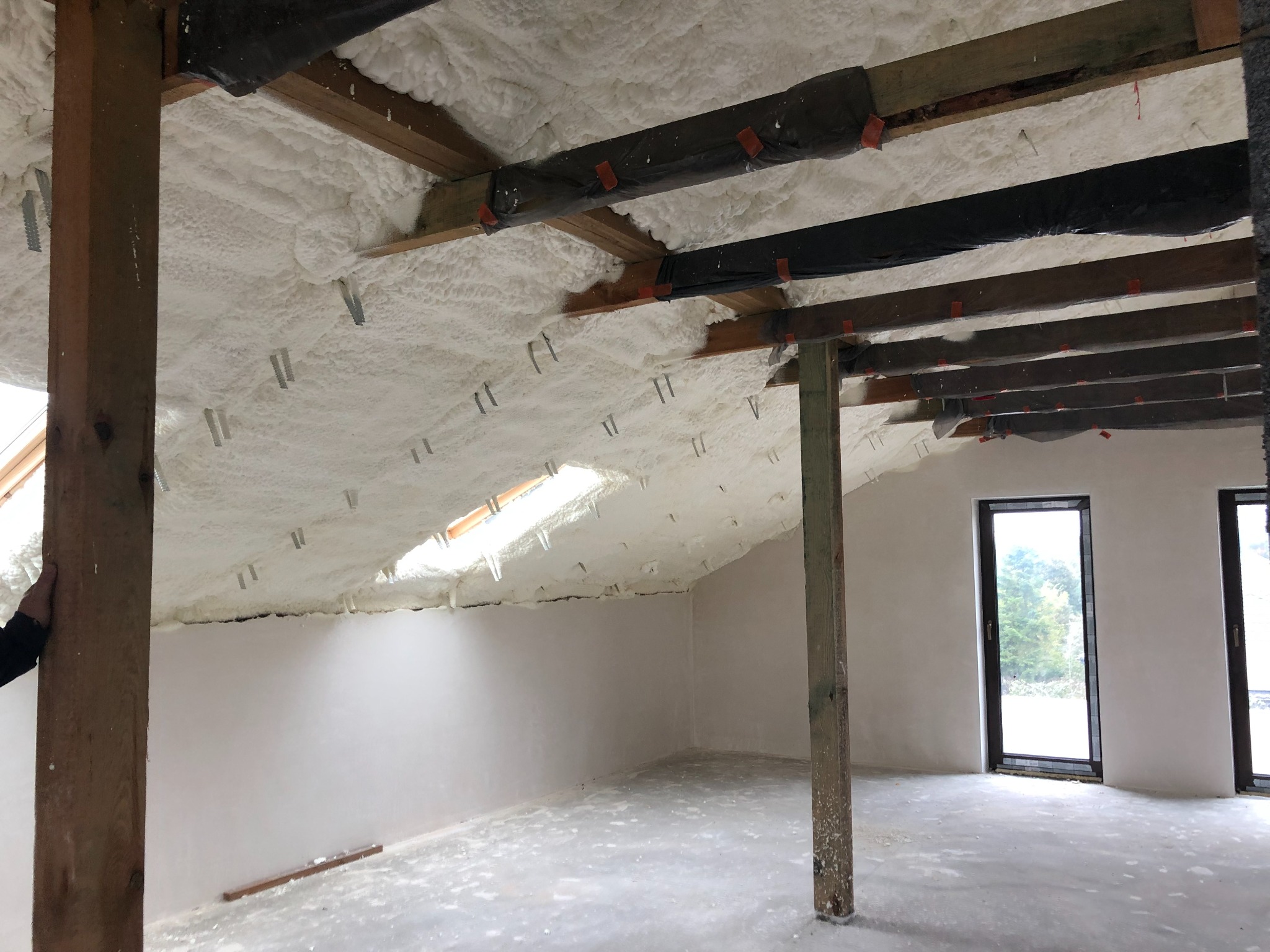 Poddasze z oknem dachowym po natrysku pianki poliuretanowej, widoczne drewniane elementy konstrukcyjne i metalowe mocowania. Ściany i podłoga przygotowane do dalszych prac wykończeniowych.