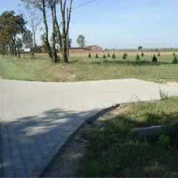 Fragment chodnika z kostki brukowej łączący drogę z polem, widoczne drzewa i krzewy, w tle budynek gospodarczy.