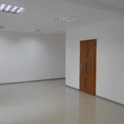 Puste, nowo wyremontowane pomieszczenie biurowe z drewnianymi drzwiami i błyszczącą podłogą z płytek, oświetlone przez rastrowe lampy sufitowe.